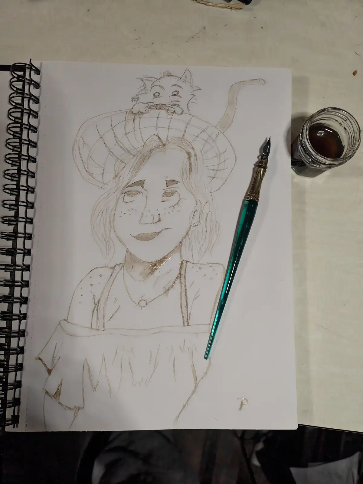 A sketch in a spiral-bound notebook shows a person with a cat perched on their head, drawn with brown ink. Beside the…