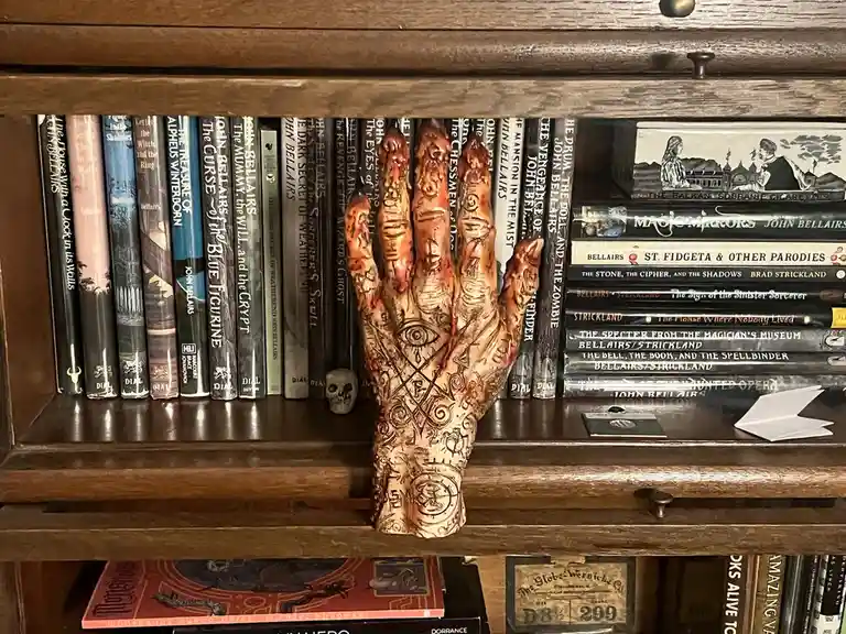 A realistic, bloody, and tattooed hand prop with occult symbols and an eye in the center is placed on a wooden bookshelf…