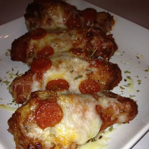 A close-up photo of four chicken wings arranged on a white plate, each covered in melted cheese and topped with pepperoni…