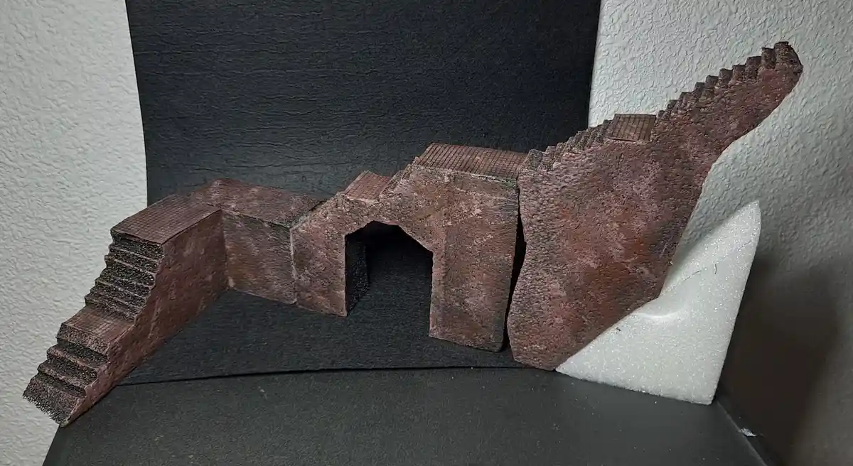 A handmade model of a castle wall with stairs and an archway, painted in a rusty brown color, is displayed on a black…