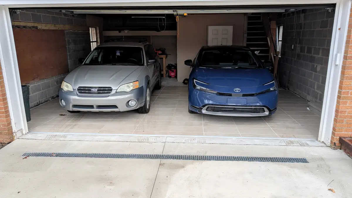 Two cars are parked side-by-side in a garage: a silver Subaru Legacy on the left and a blue Toyota Prius Prime on the right…