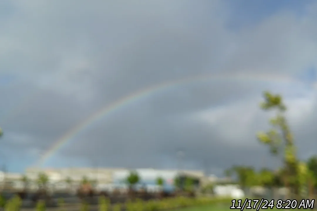 A blurry outdoor photo shows a faint rainbow arcing across a cloudy sky above indistinct buildings and trees. The timestamp…