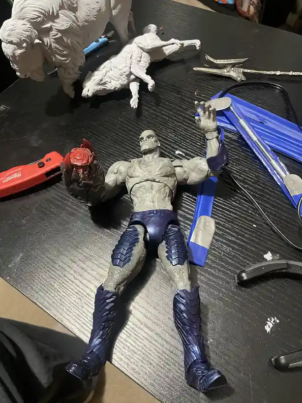A partially painted action figure, resembling a muscular, grey-skinned character with blue armor, lies on a workbench…