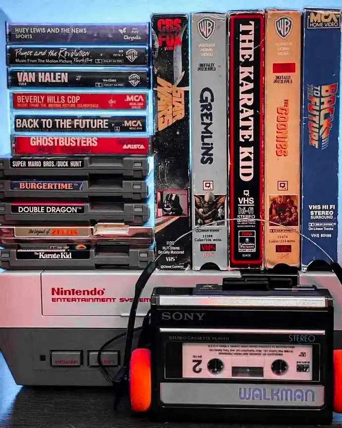 A photo shows a stack of VHS tapes and game cartridges arranged vertically behind a vintage Sony Walkman and a Nintendo…