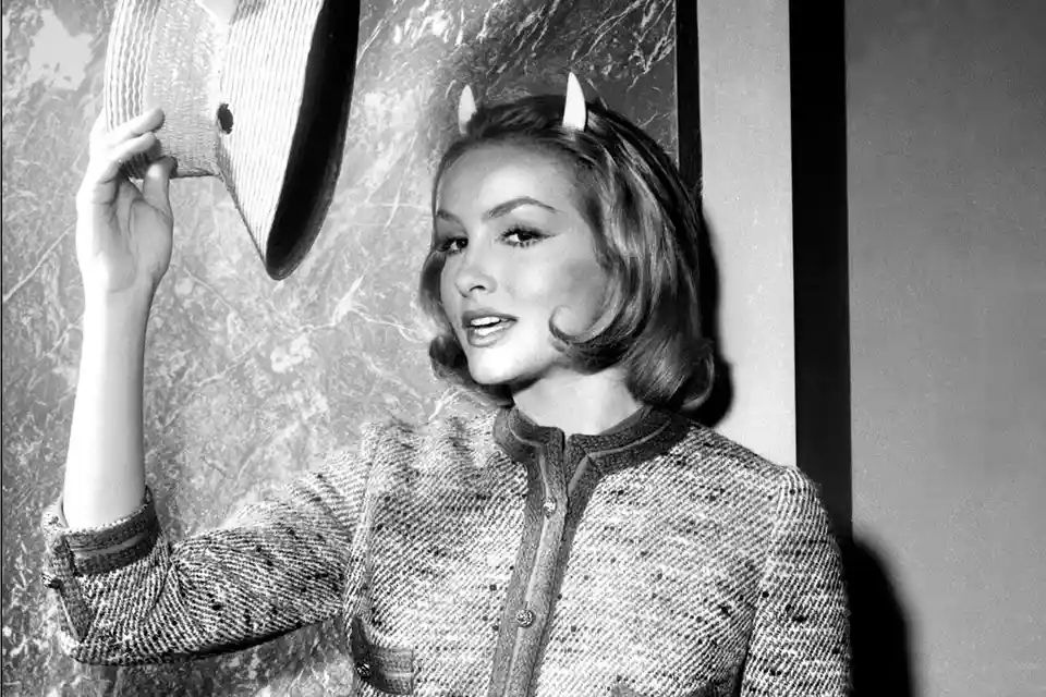 A black-and-white photo of a woman with small white devil horns on her head, holding a hat up near her face. She wears a…