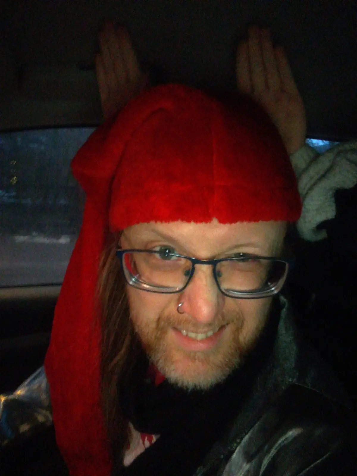 A person with glasses and a nose ring wears a red Santa hat with a long, floppy red hood, smiling at the camera while seated…