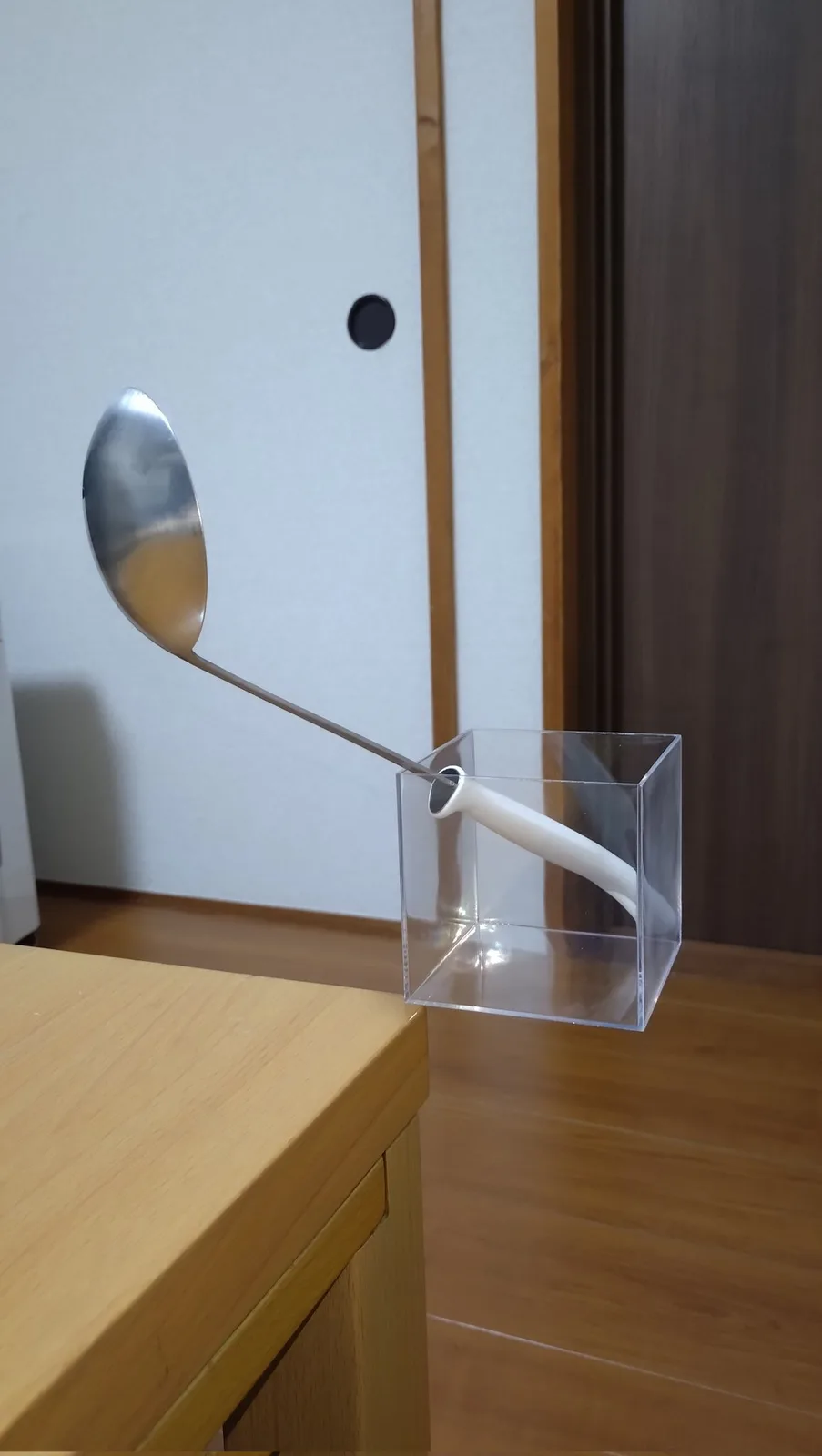 A metal spoon with a white handle is balanced upright inside a clear plastic cube, resting on a wooden table. The spoon's…