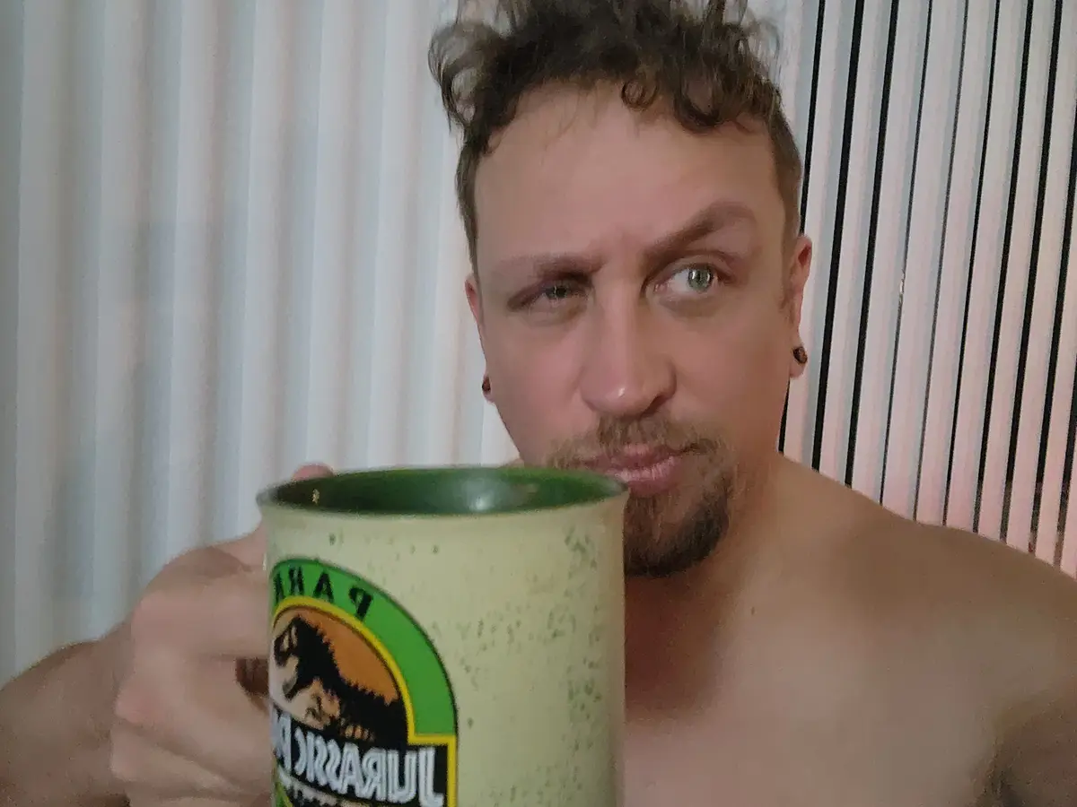 A shirtless man with curly hair and facial hair holds a green Jurassic Park mug up to his face, obscuring his mouth and…