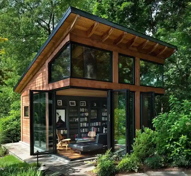 A modern wooden cabin nestled in lush green trees, featuring large glass windows and doors revealing a cozy interior with…
