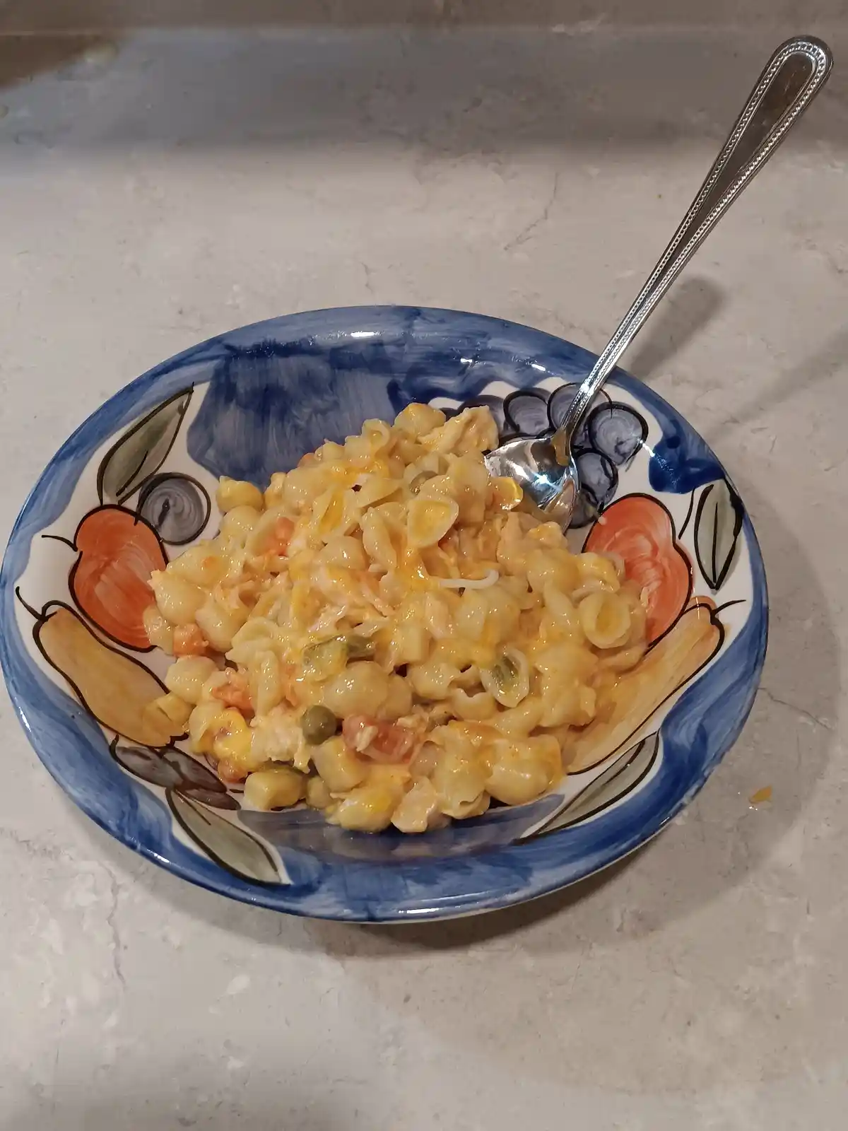 A bowl of macaroni and cheese with a creamy orange sauce, capers, and diced tomatoes sits in a decorative ceramic bowl with…
