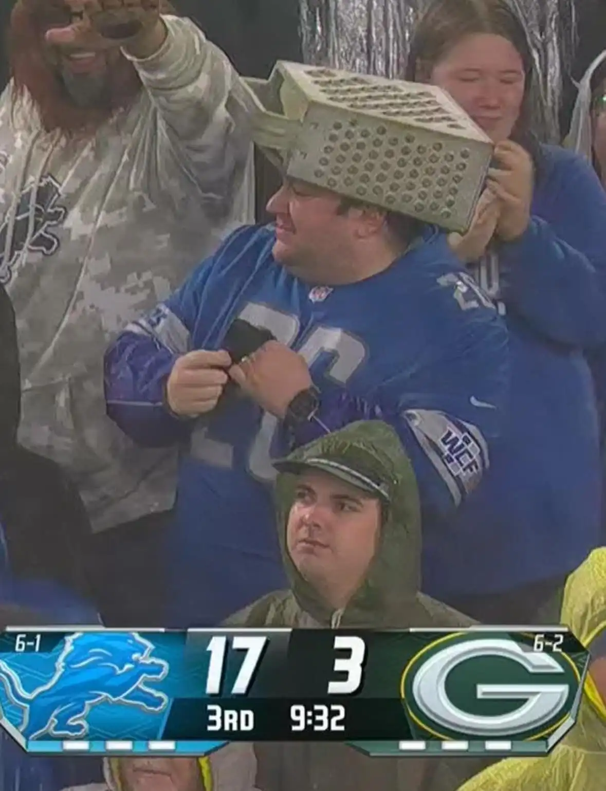 A man in a blue Detroit Lions jersey has a metal grater placed on his head by a woman behind him while he looks down…