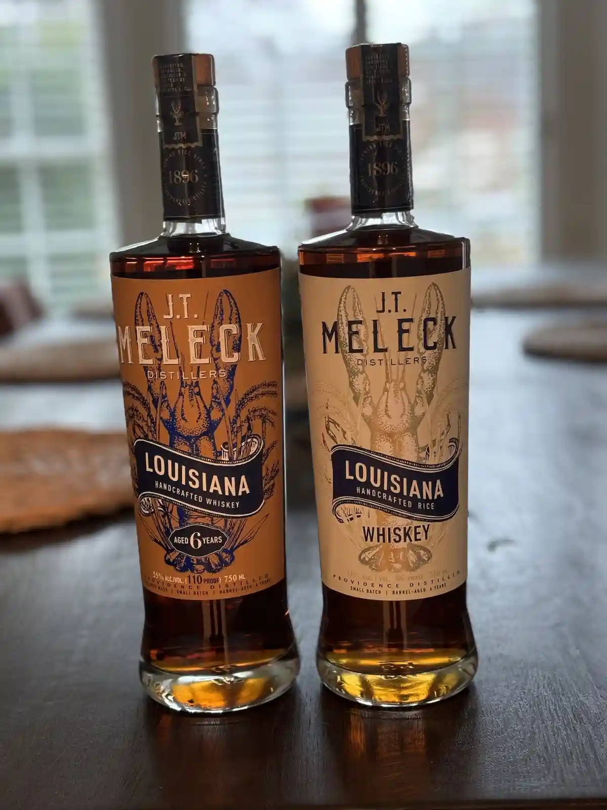 Two bottles of J.T. Meleck Distillers' Louisiana whiskey sit side by side on a wooden table; the left bottle is labeled…