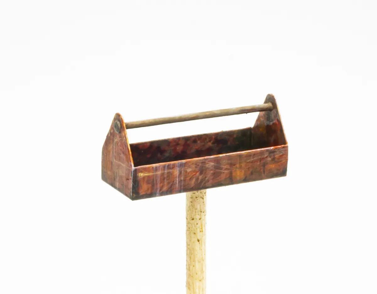 A small, weathered wooden toolbox with a metal handle is mounted on the head of a single wooden matchstick against a plain…