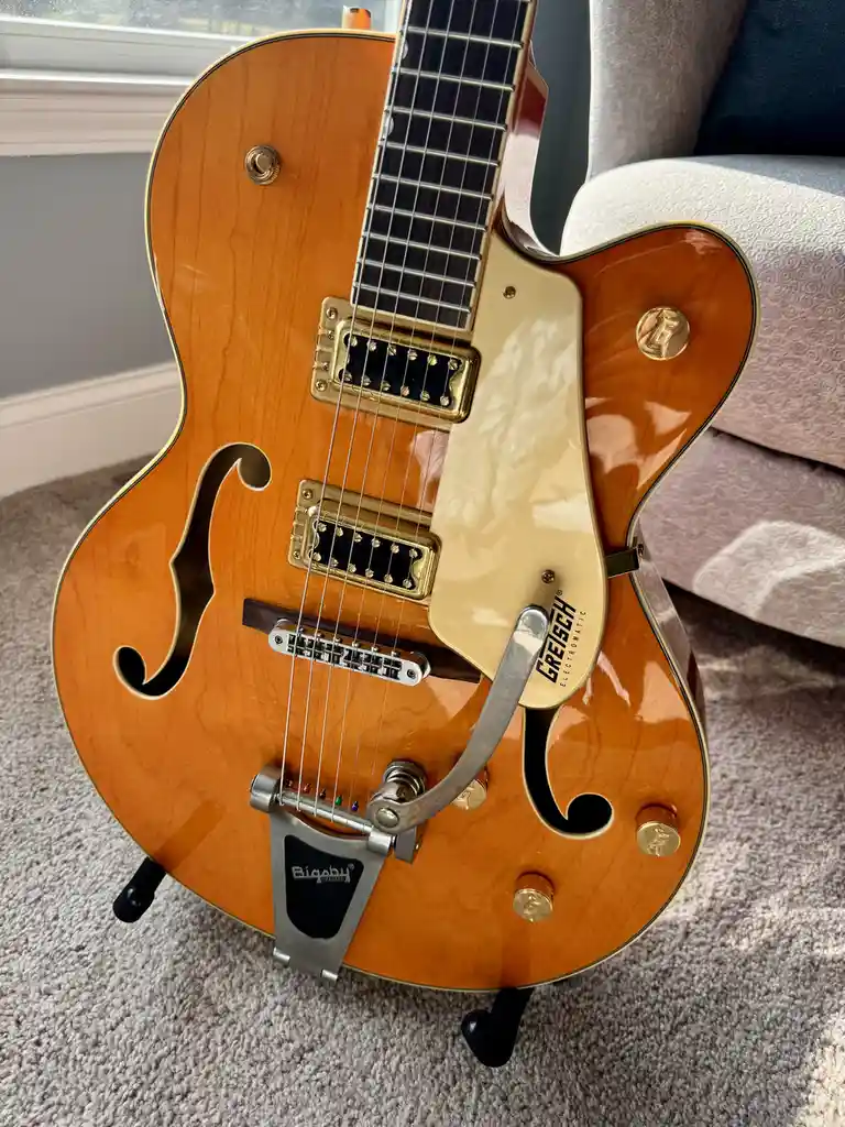 A close-up photo of a sunburst-finish Gretsch Electromatic hollow-body electric guitar resting on a carpeted floor, with a…
