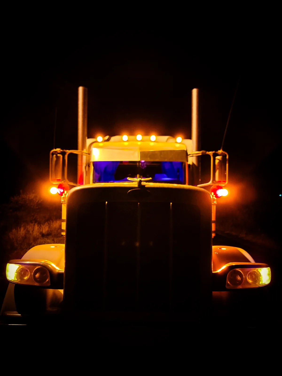 A front-facing photo of a large white semi-truck at night, its headlights and roof lights glowing orange against a black…
