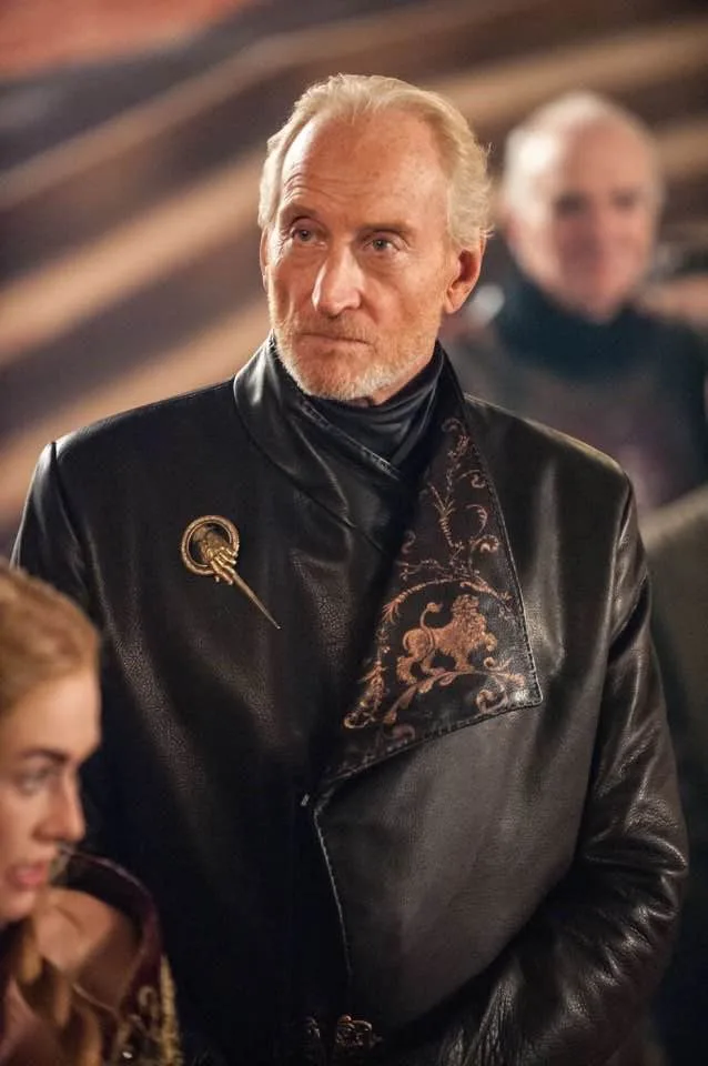 A man with white hair and a beard, wearing a black leather jacket with a lion emblem and a pin, looks sternly at the camera…