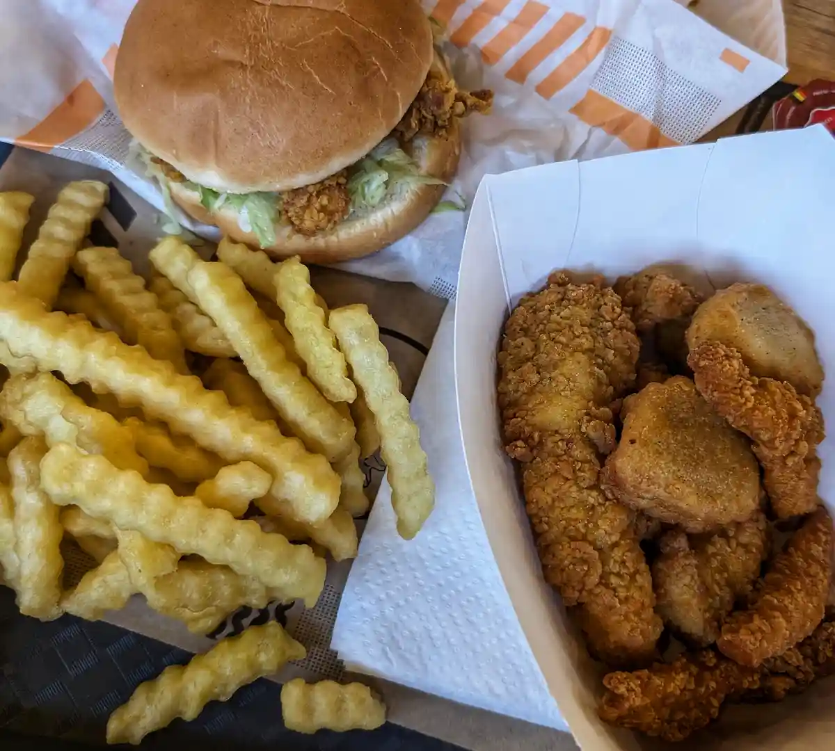 A close-up photo shows a fried chicken sandwich on a bun with lettuce, next to a white paper container filled with fried…