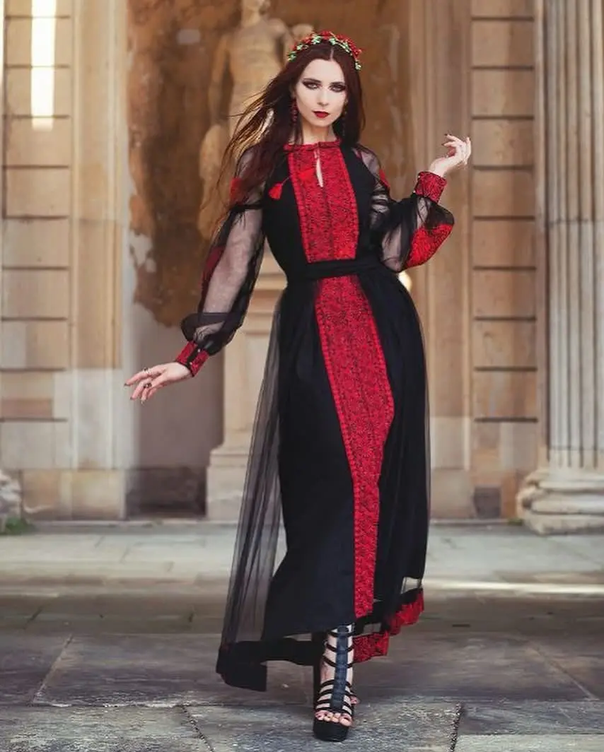 A woman with long dark hair and dramatic makeup stands in a classical stone courtyard, wearing a striking black and red…