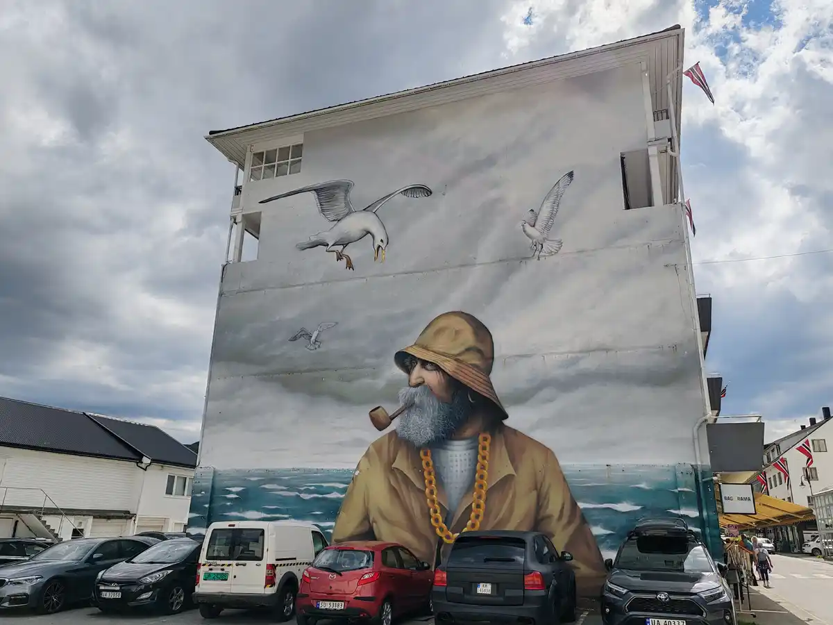 A large mural on a building depicts a bearded man in a hat smoking a pipe, with several seagulls flying around him, one…