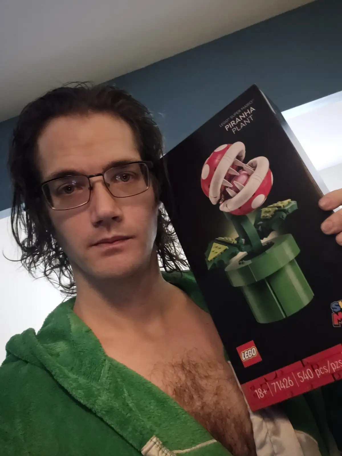 A man with glasses and a green towel draped over his shoulders holds up a LEGO Super Mario Piranha Plant set box. The box…