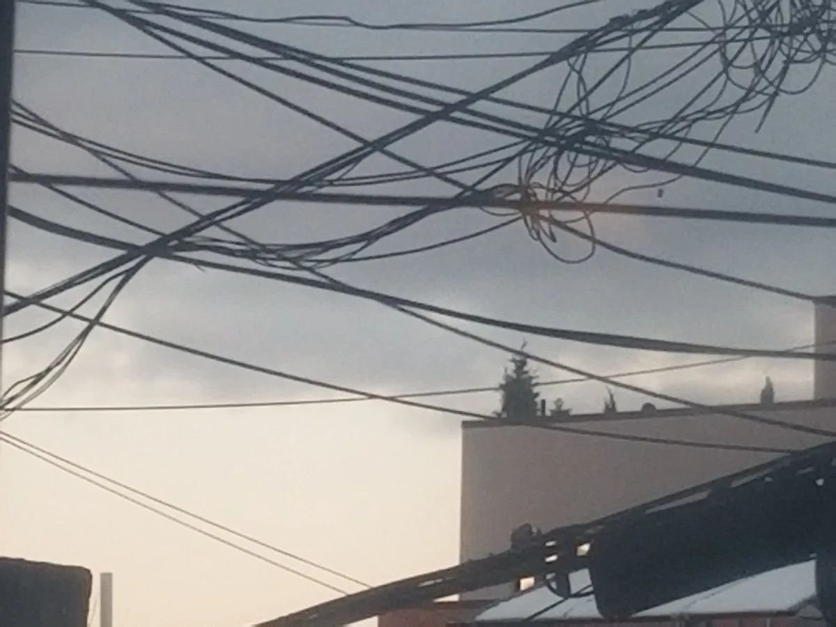 A photograph shows a dense, chaotic tangle of overhead power lines against a cloudy sky, with the silhouette of a building…