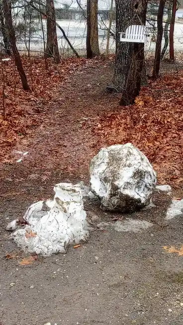 A snowy outdoor scene shows a large, partially melted snowball and a smaller, melted snow pile on a dirt path covered in…