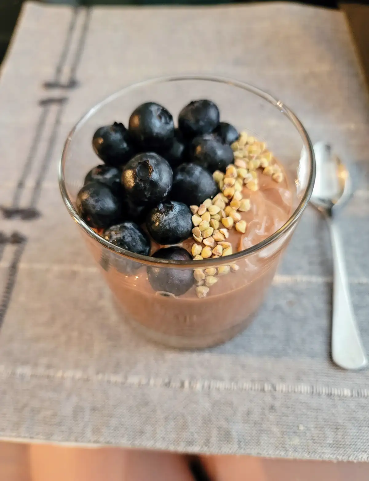 A clear glass bowl holds chocolate pudding topped with a pile of fresh blueberries and a line of buckwheat groats, sitting…