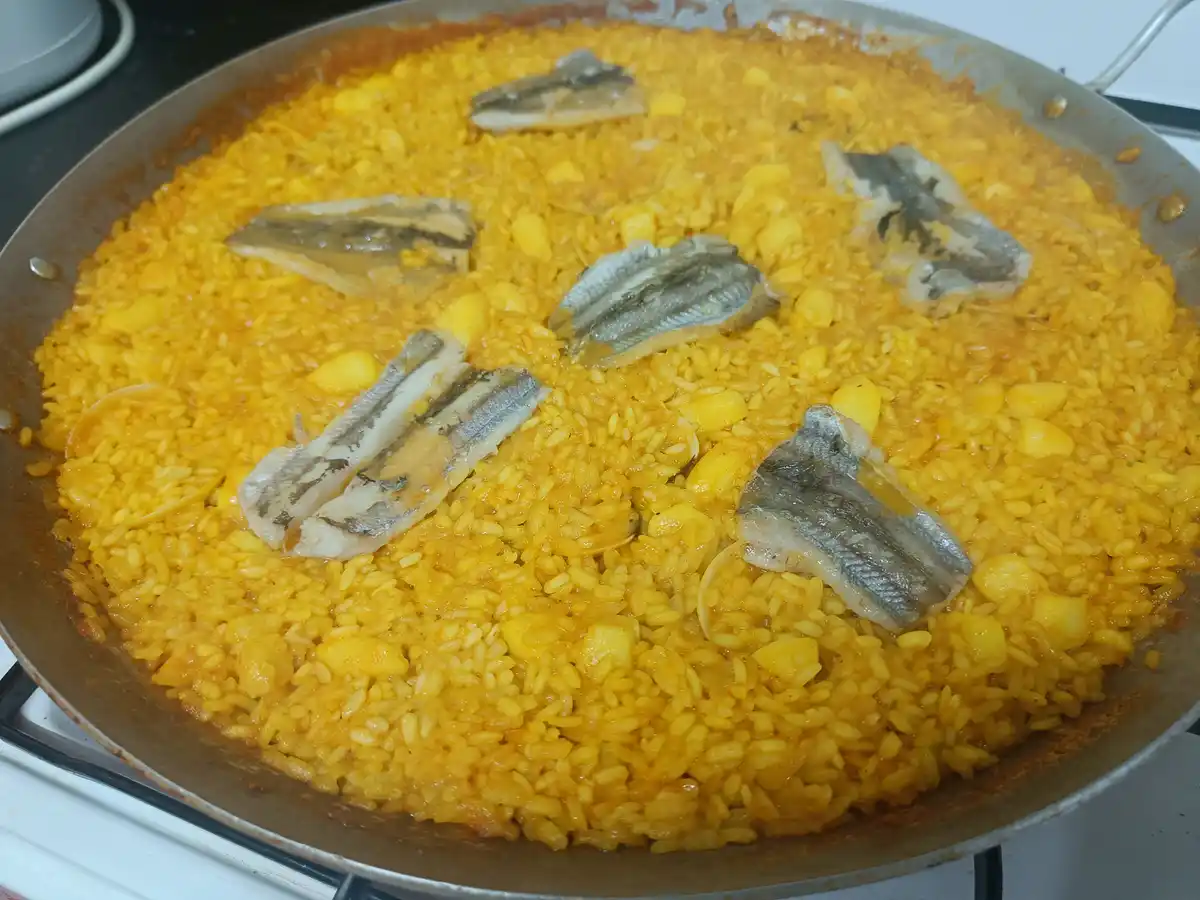 A close-up photo shows a large, shallow pan filled with golden-yellow paella rice, mixed with chunks of potato and topped…