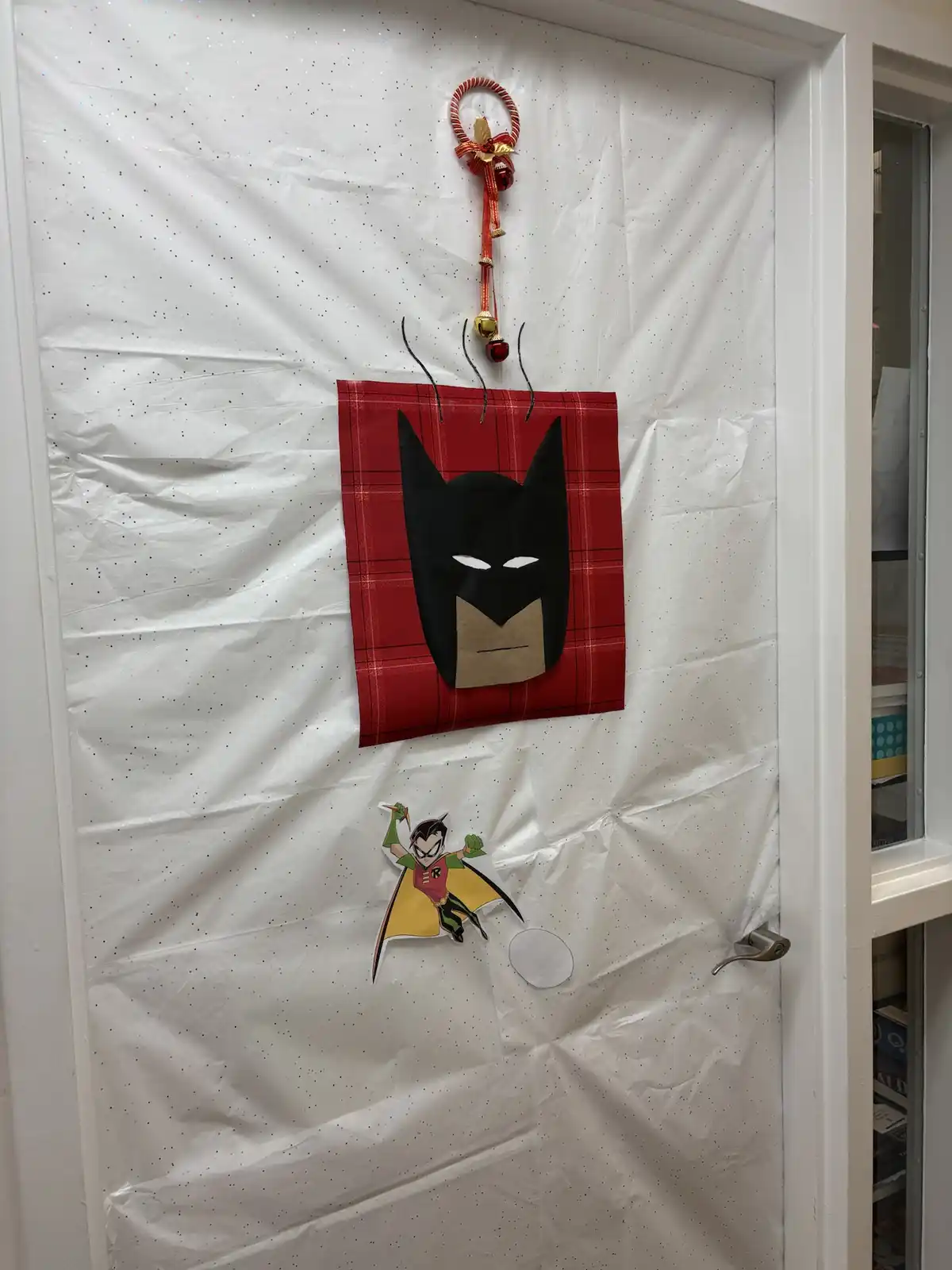 A door decorated with white glitter paper features a handmade Batman head emerging from a red plaid mug, with steam lines…