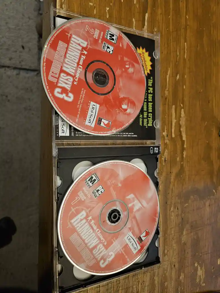 Two discs from the PC game 'Tom Clancy's Rainbow Six 3: Raven Shield' are shown in their cases on a wooden surface. The top…