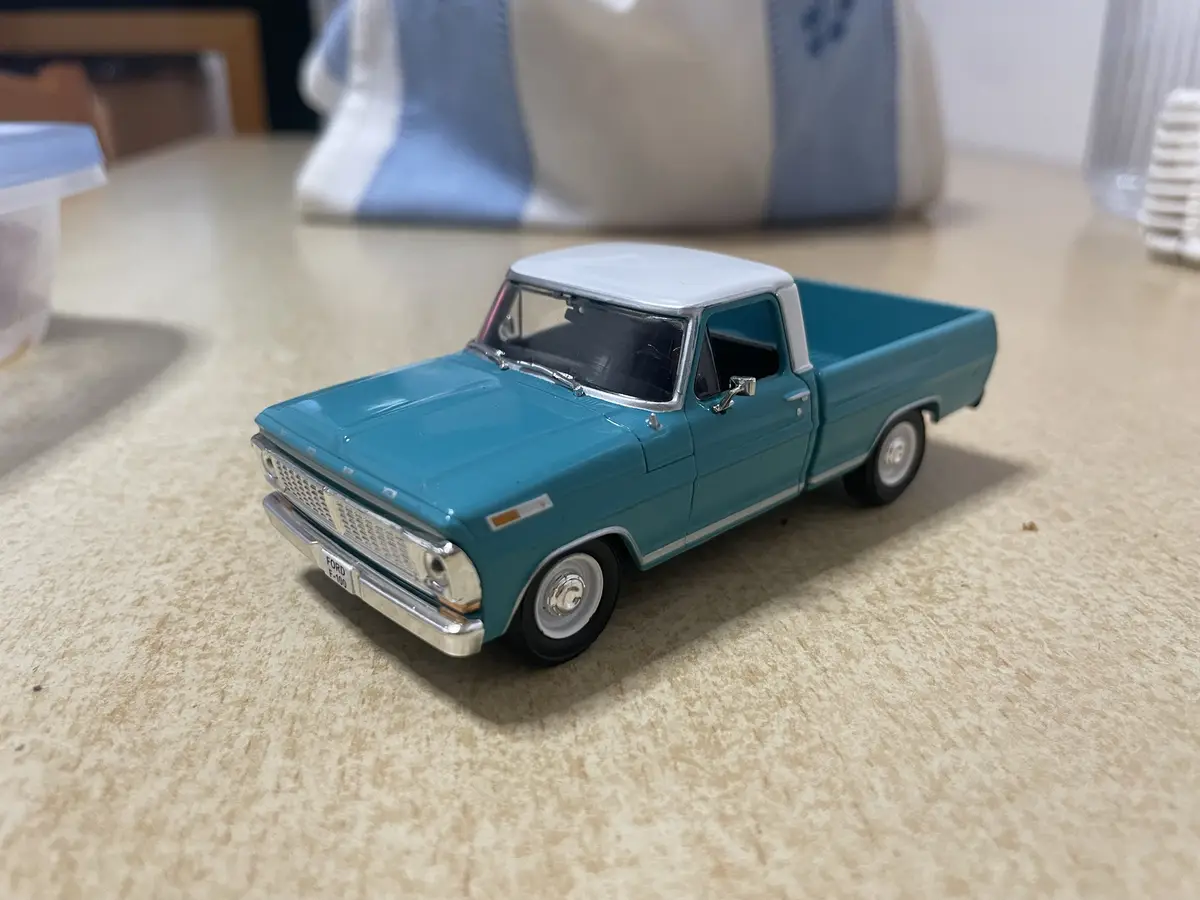 A teal and white die-cast model of a Ford F-100 pickup truck sits on a speckled countertop, with household items like a…