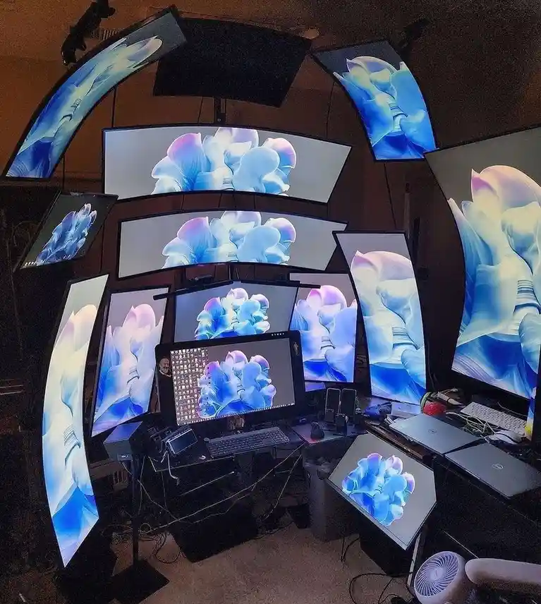 A room filled with numerous computer monitors arranged in a curved, multi-tiered setup around a central desk, all displaying…