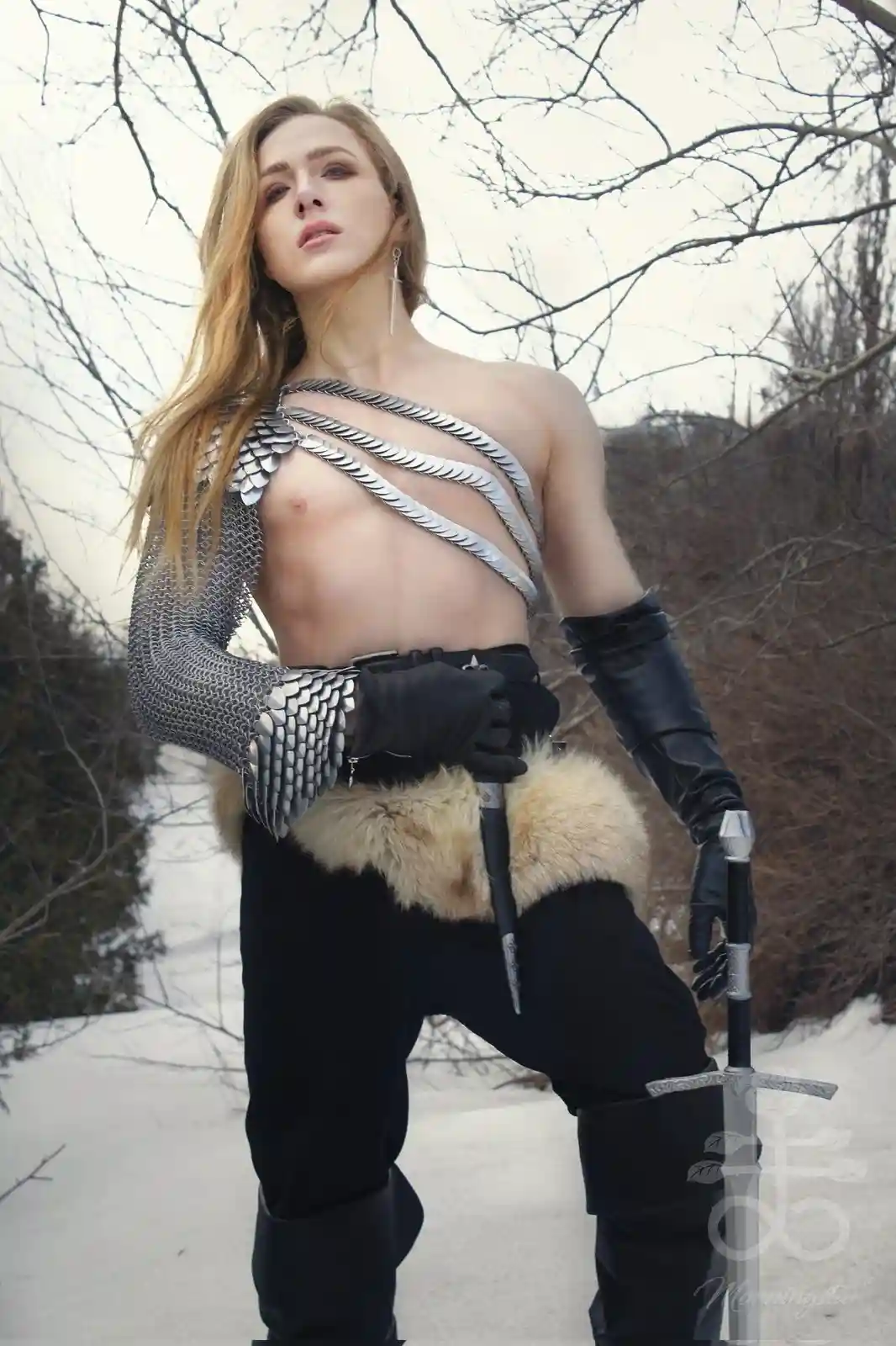 A person with long blonde hair stands in a snowy, wooded area wearing partial medieval armor including chainmail on one arm…