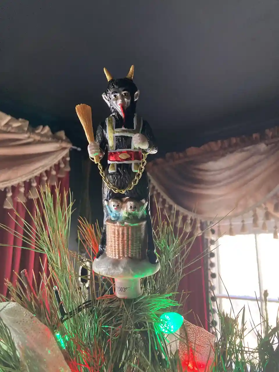 A close-up photo shows a Christmas tree topper shaped like a demonic figure with horns, pointed ears, and a red tongue…