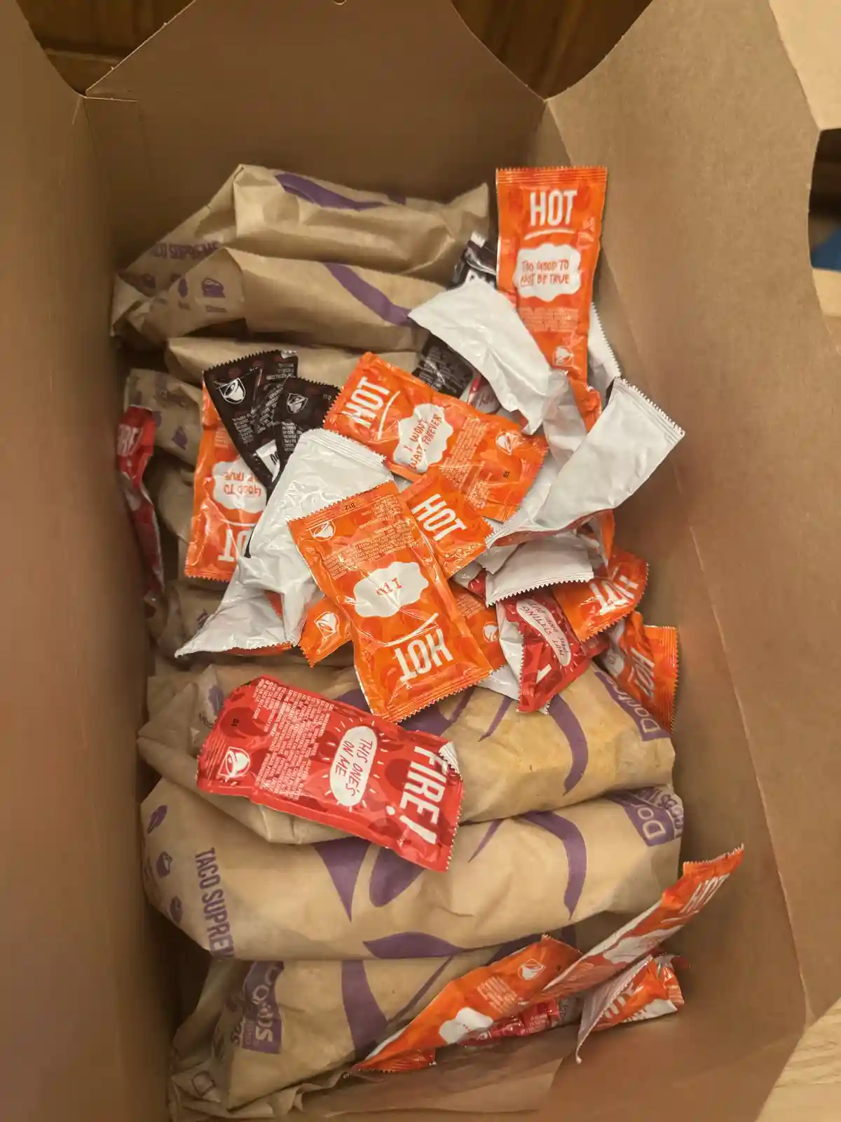 A cardboard box overflowing with Domino's Taco Supreme pizza bags and a large pile of hot sauce packets in orange, red, and…