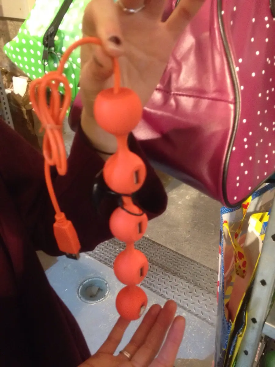 A person holds up a bright orange, USB-powered, multi-bulb sex toy with a coiled cable, while standing near a pink polka-dot…