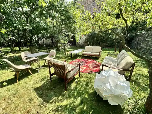 An outdoor garden scene with lush green grass, trees, and a stone wall, featuring mid-century modern furniture including…