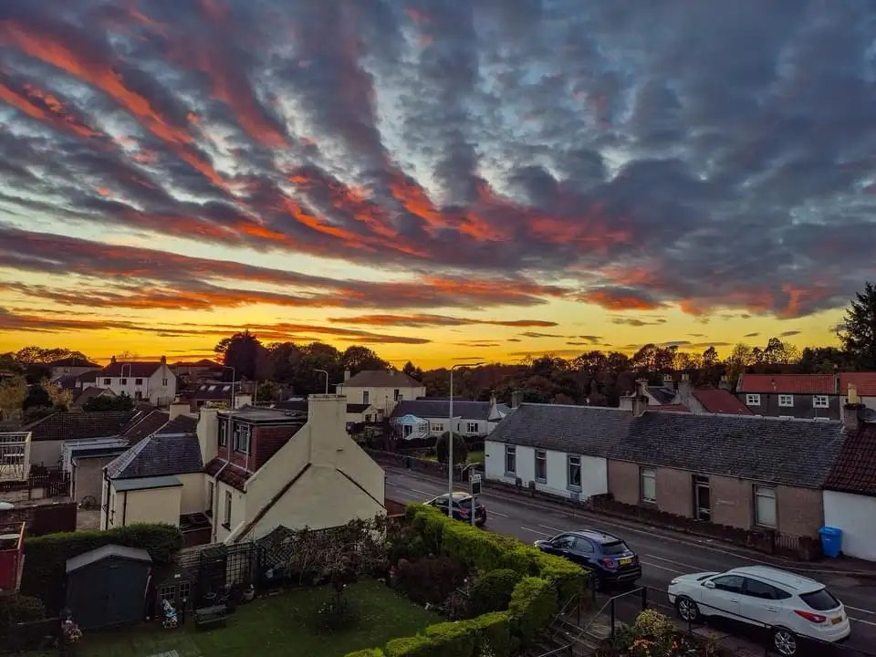 A scenic photograph of a residential street at sunset, featuring houses with tiled roofs, parked cars, and vibrant orange…