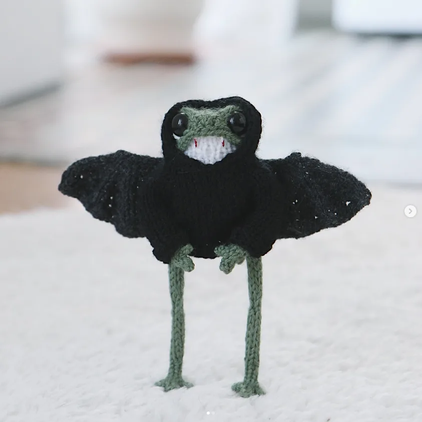 A small, hand-knitted plush bat with black wings, green limbs, and a white mouth with red stitches for teeth stands on a…