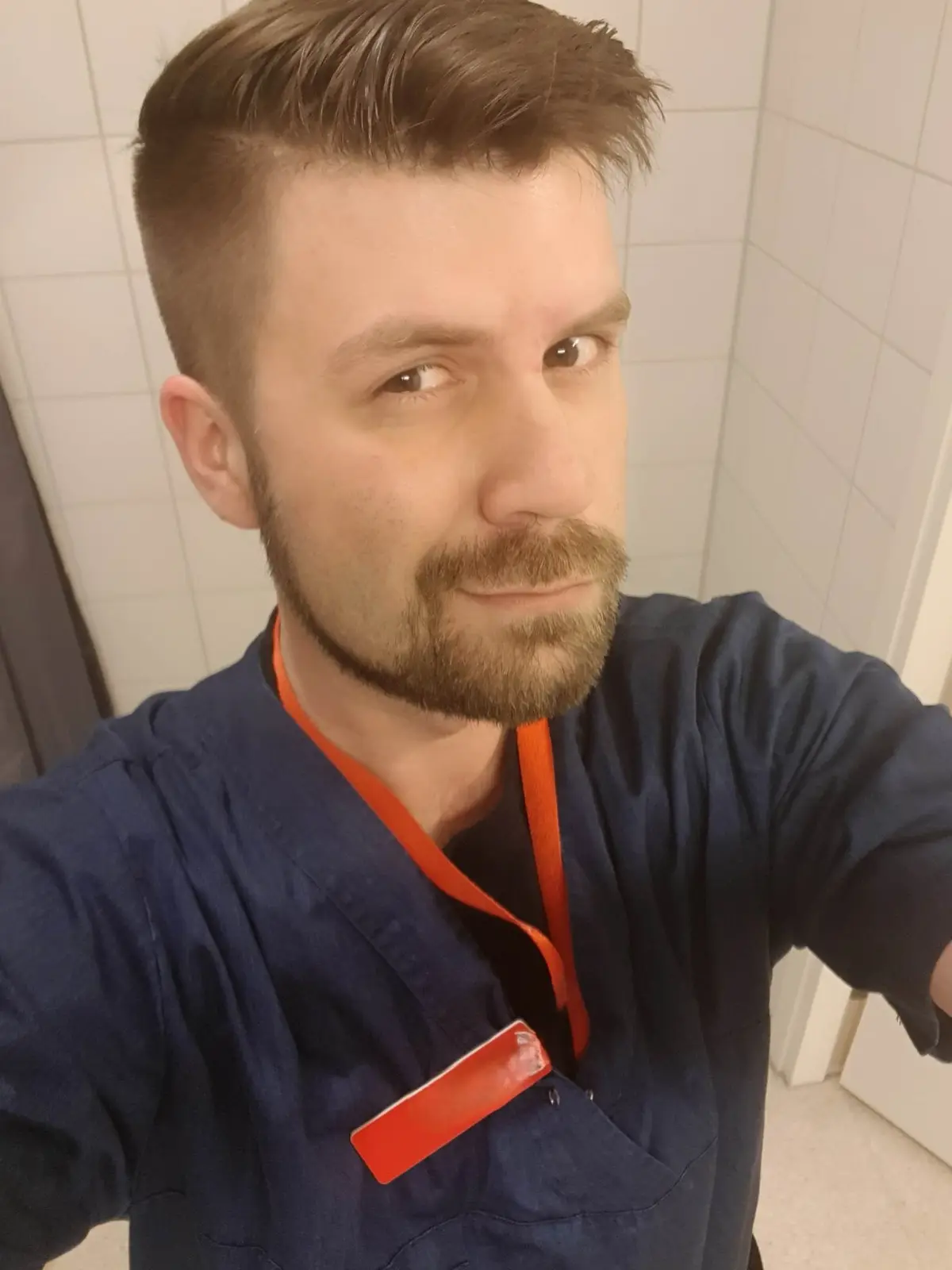 A man in blue medical scrubs with an orange lanyard and a red ID badge takes a selfie in a tiled room, looking directly at…