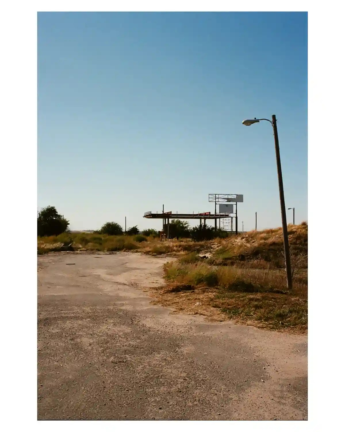A desolate, cracked asphalt road leads toward a dilapidated, empty billboard structure under a clear blue sky, with dry…
