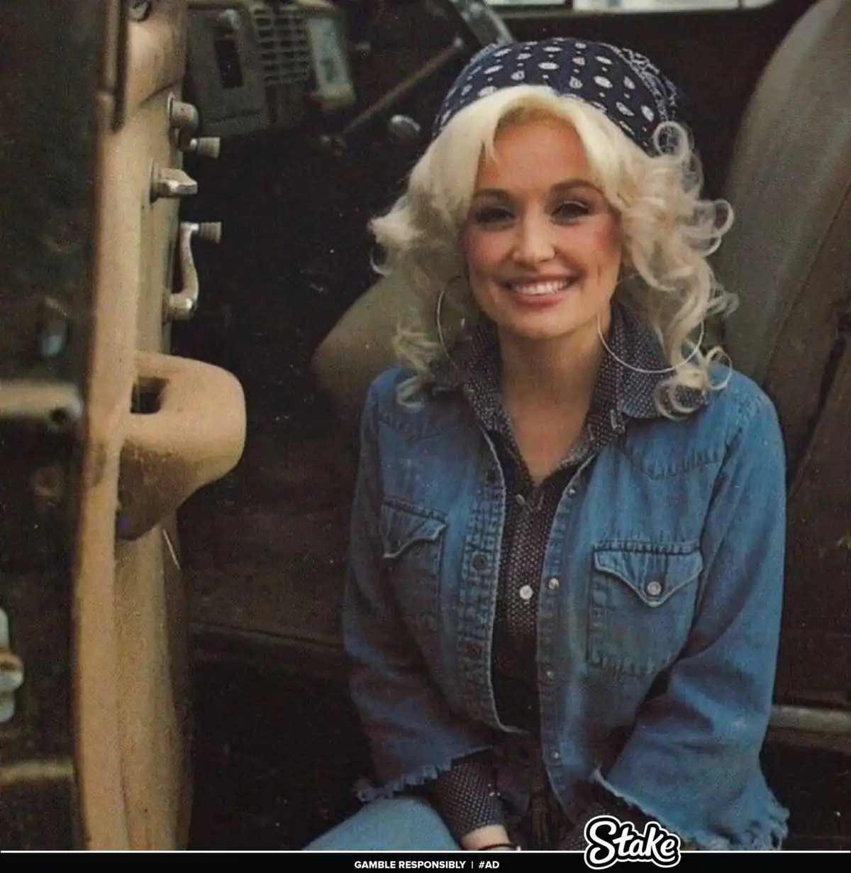 A woman with blonde curly hair, wearing a blue bandana, large hoop earrings, and a denim jacket over a patterned shirt…