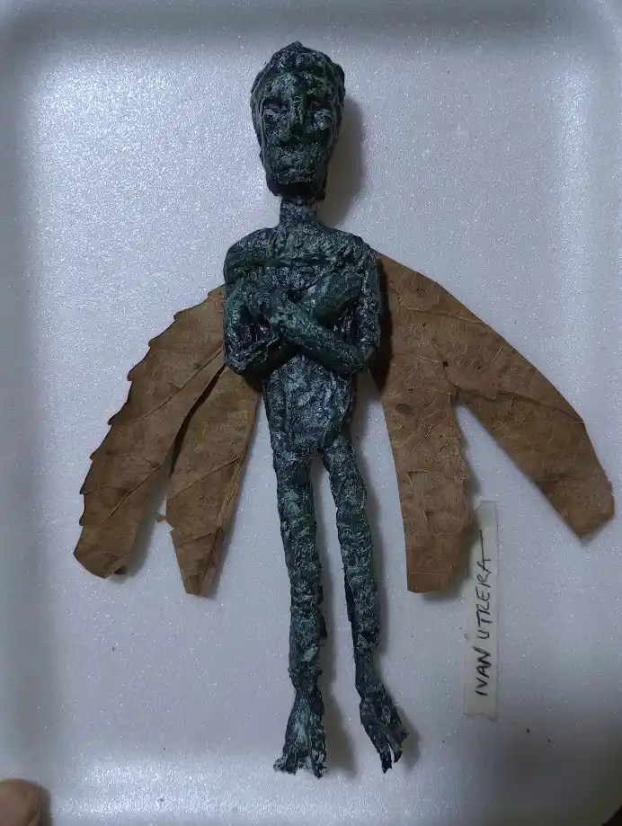 A small, greenish-brown figurine with a humanoid shape and leaf-like wings is displayed on a white surface. A handwritten…