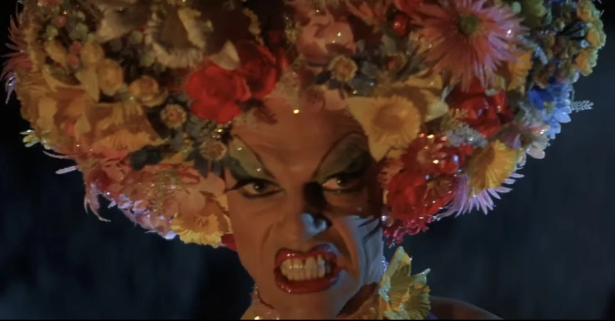 A person with dramatic makeup and a large, colorful floral headdress stares intensely with a grimace and bared teeth. The…