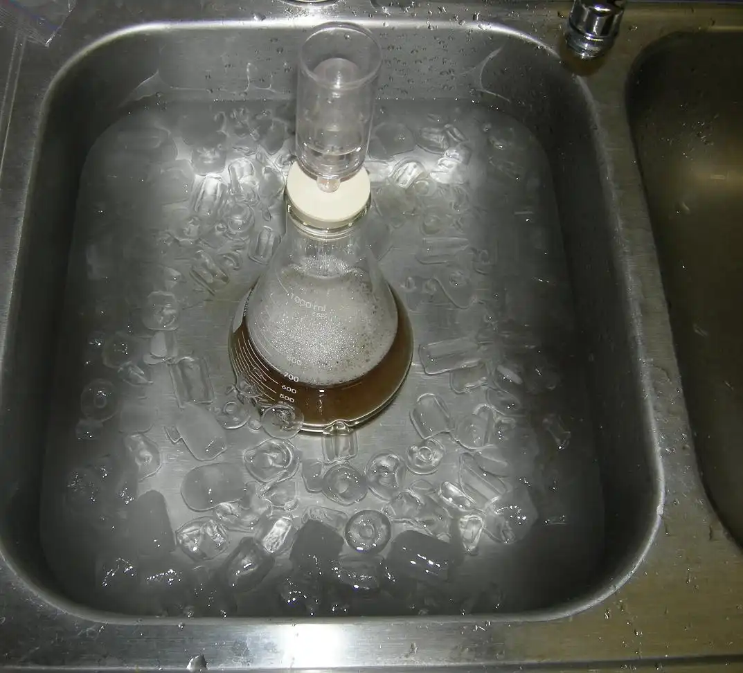 A conical flask containing a brown liquid and foam sits in a metal sink filled with ice and water, with a glass tube…