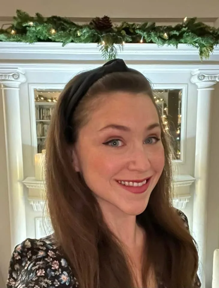 A woman with long brown hair and a black headband smiles at the camera, wearing a floral top. Behind her is a white…