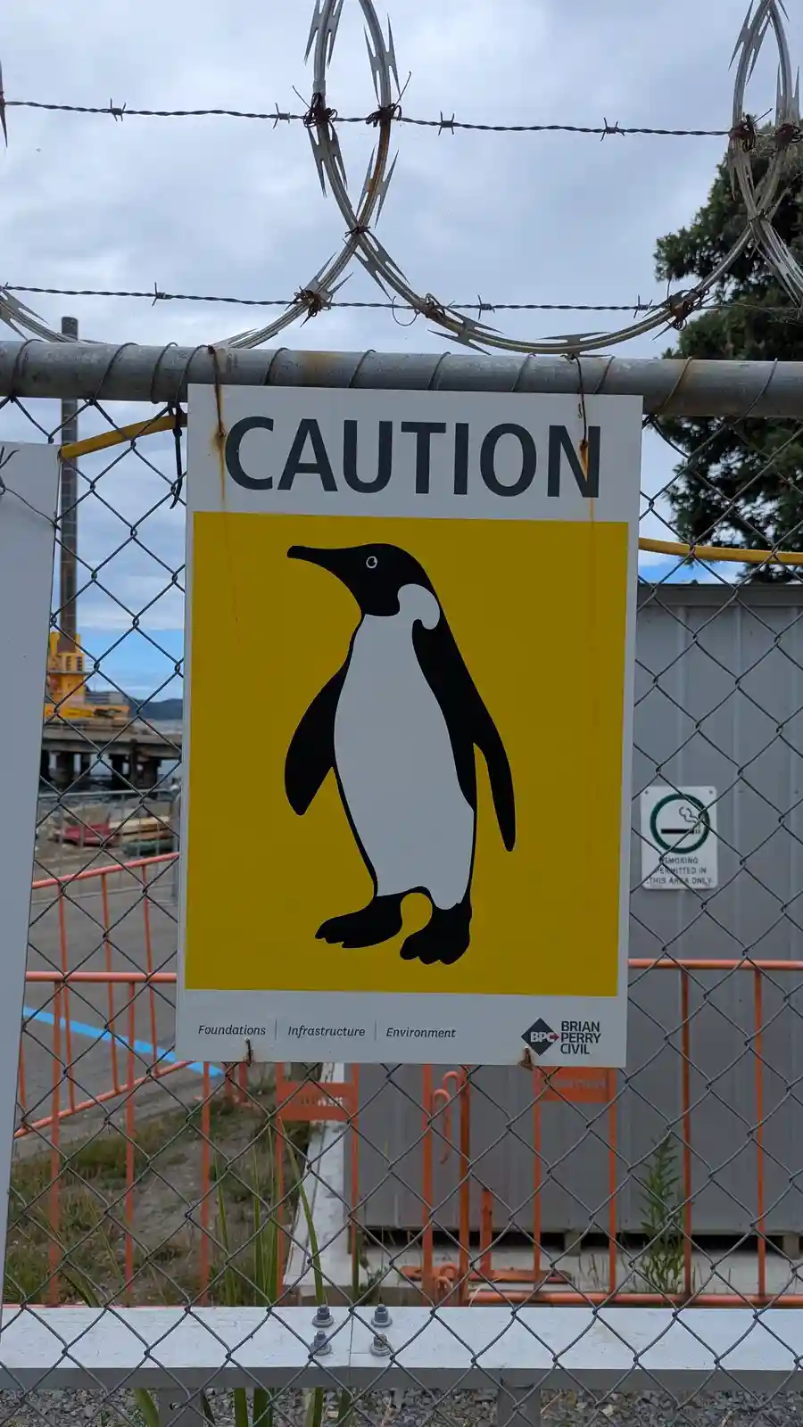 A yellow and white caution sign featuring a black-and-white penguin silhouette is attached to a chain-link fence topped with…