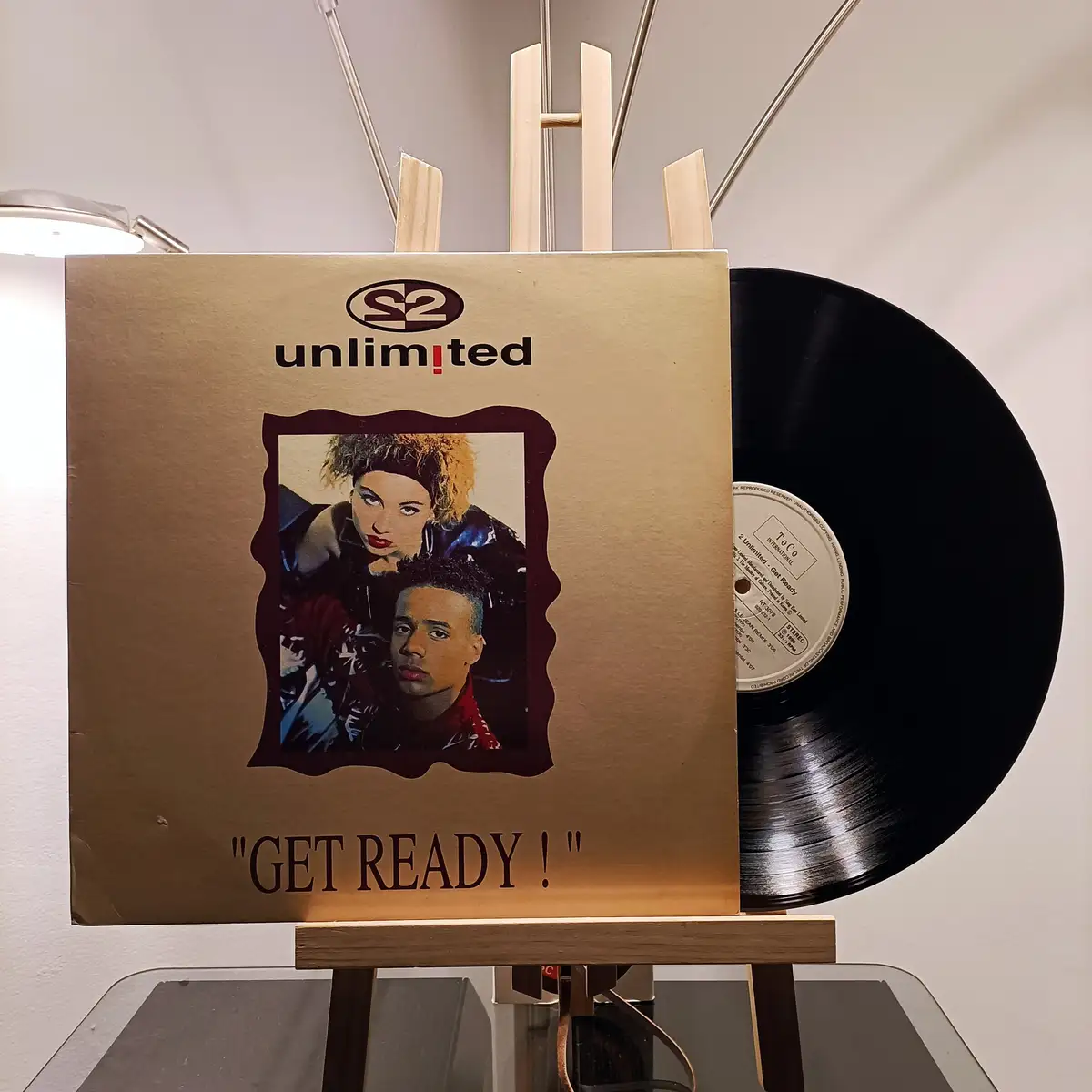 A gold-colored vinyl record sleeve for the group S2 Unlimited, featuring a photo of two people, is displayed on a wooden…