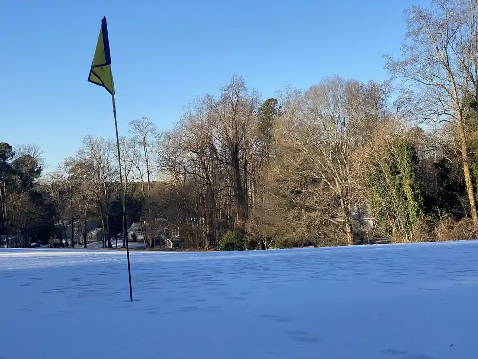 A bright yellow golf flag stands in a snow-covered field under a clear blue sky, with bare trees and houses visible in the…