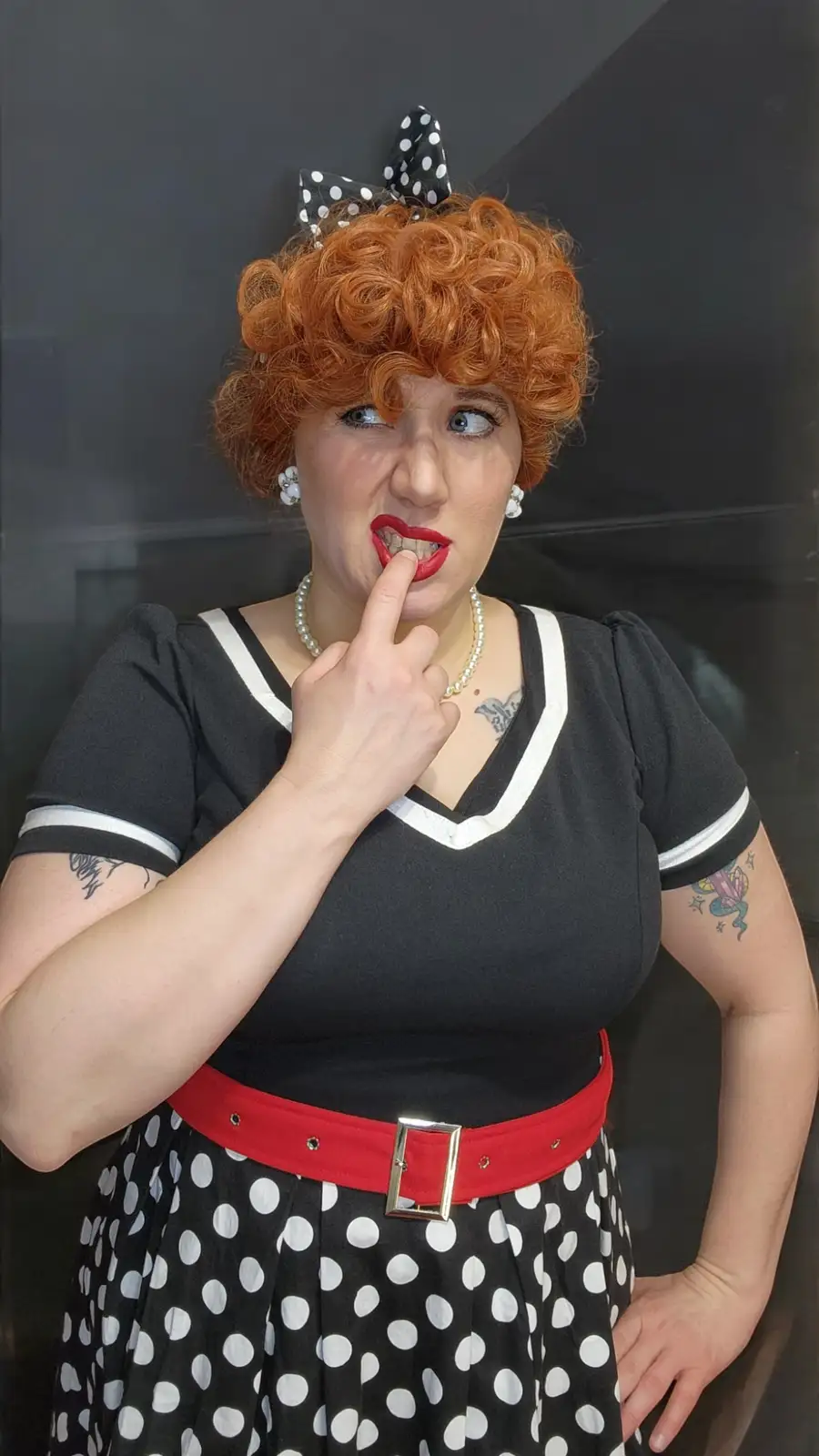 A person with curly red hair and a black-and-white polka dot dress with a red belt poses with a mischievous expression…